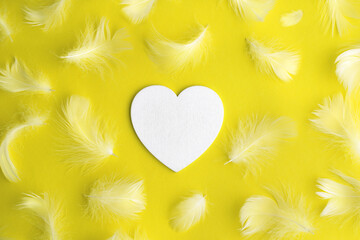 Heart and soft fluffy white feathers on bright yellow paper background. Top view. Place for...