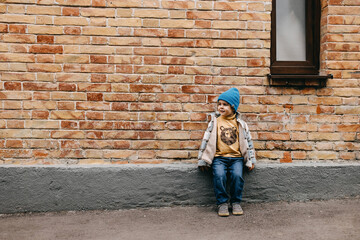 Toddler boy outdoors sitting by a brick wall, on city streets.