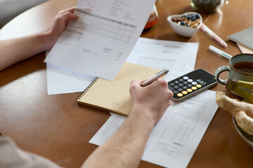 Cropped shot of unrecognizable man checking documents utility bills taking care of personal finance and calculating expenses on a smartphone making notes in notebook at a kitchen table indoors.