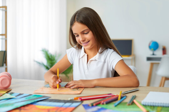 Smiling cute girl drawing, painting picture with interest using colorful pencils sitting at table at home. Preteen girl in white polo shirt looking at picture. Education, art, homeschooling concept.