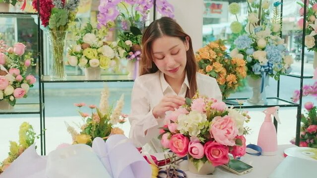 One young beautiful Asian female florist worker arranging and decorating bunch of fresh blossoms, valentine orders, smiling with happy work in colorful slower shop, small business SME entrepreneur. 