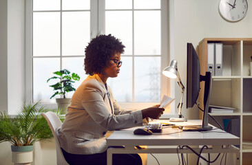 Side view portrait of a concentrated serious african american business woman in glasses sitting at the desk on her workplace working on a laptop and looking at documents at office.