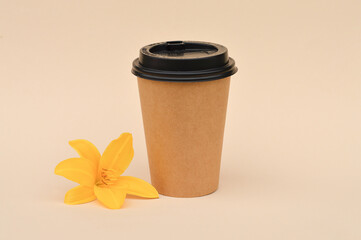 A cardboard cup for coffee, suitable for reuse, next to a yellow lily flower on a beige background,...