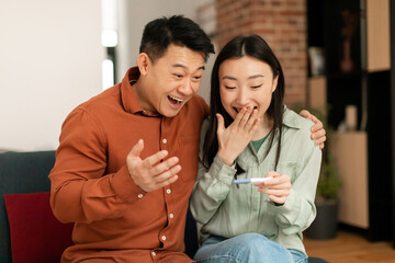 Fototapeta na wymiar Happy japanese middle aged husband and his young wife rejoicing positive pregnancy test, sitting in living room interior