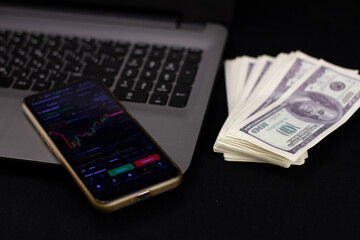 On a black background, a laptop keyboard with a cryptocurrency market open on the phone and dollar banknotes. Buying and selling cryptocurrencies on the stock exchange on the Internet.