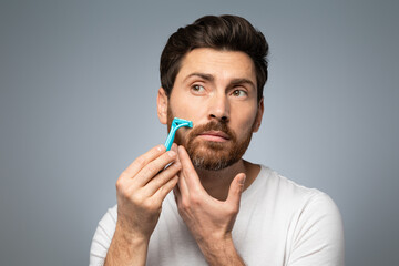 Handsome bearded middle aged man in t-shirt shaving his beard while standing over grey studio background