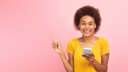 Laughing shocked young black curly lady with smartphone pointing her finger at empty space