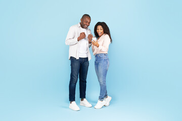Overjoyed black couple holding smartphone and shaking fists in joy and excitement, blue studio background