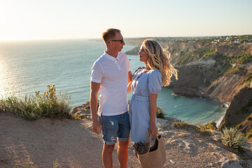 The romantic couple is looking at the sunset and hugging. Travellers admire the beautiful landscape of nature, water, sun rocks