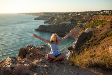 The traveller's girl with her hands raised sit and admires the nature of the rocks, sun and water. the tourist woman is dressed in summer clothes, blue sundress.