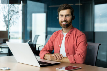 Portrait of mature businessman in red shirt inside office, man with beard and video call headset smiling and looking at camera, online customer support worker with laptop. - Powered by Adobe
