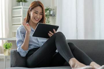 A pretty smiling Asian businesswoman sitting happily holding a tablet on the sofa looking at the desired product details in an online shopping cart in the office.