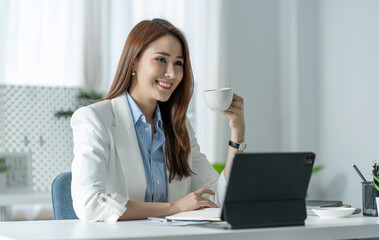Pretty smiling Asian businesswoman sitting with a cup of coffee relaxing on the desk after confirming financial documents Happy validation at the office.