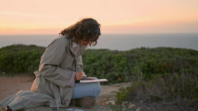 Inspired girl painting evening ocean view. Focused artist relaxing rocky hill