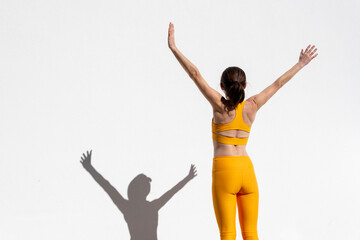 Rear view of a fit, sporty woman with her arms raised standing by a white wall, fitness concept.