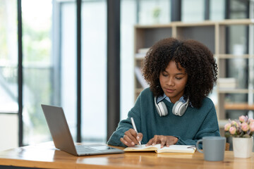 Cute african american female student wearing headphones with afro dreadlocks, studying remotely from home, using a laptop, and taking notes on a notepad online.