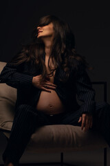 Naklejka premium Beautiful pregnant girl at the studio. A pregnant girl in a man's suit.Stunning pregnant woman - maternity photography of a beautiful young expecting mother to be on a black background, studio photogr