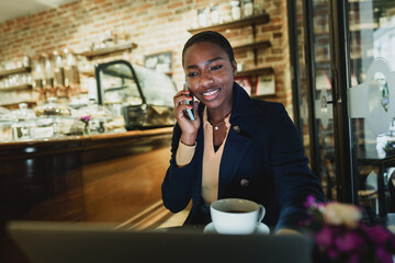 Portrait of a smiling young woman using laptop and talking on mobile phone in coffeeshop