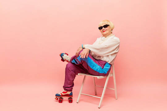 Sportive vibe. Beautiful old woman, grandmother in stylish sportive trousers posing on vintage rollers over pink studio background. Concept of age, fashion, lifestyle, emotions, facial expression