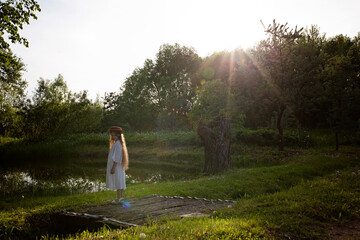 Pond. Sun. A little girl in a hat stands on the bridge. Little girl on the background of evening summer nature. Holidays outside the city.