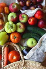 Straw bag and reusable fabric bags filled with various healthy fruit and vegetables. Wooden background, top view.