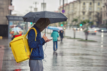 Delivery boy with smartphone and ubmrella works during rain. Delivery man with yellow backpack delivering food to costumers in rainy day. Man deliver online orders to customer. Food courier
