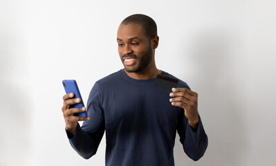 Young African-American man smiling in a casual T-shirt, posing isolated on a white background on the wall.People's lifestyle concept.Use mobile phone to make online purchases with the bank credit card
