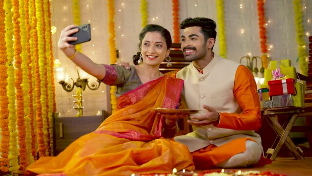 Happy smiling indian couple taking selfie on mobile phone by holding flowers plate during diwali festival celebration at home - concept of togetherness, social media sharing and traditional culture.