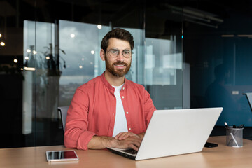 Close-up portrait of mature businessman inside office, man with beard and red shirt smiling and looking at camera, programmer developer coding software at workplace with laptop.