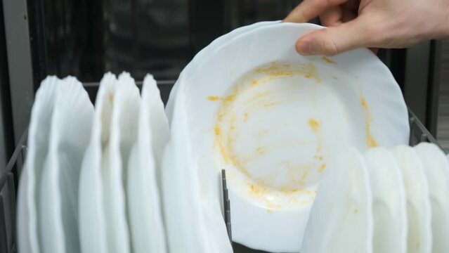 Man puts white dirty dishes in down basket of dishwasher on the kitchen. Household duties washing dishes using electrical appliances. Man puts plates to dishwasher and closes and runs it.