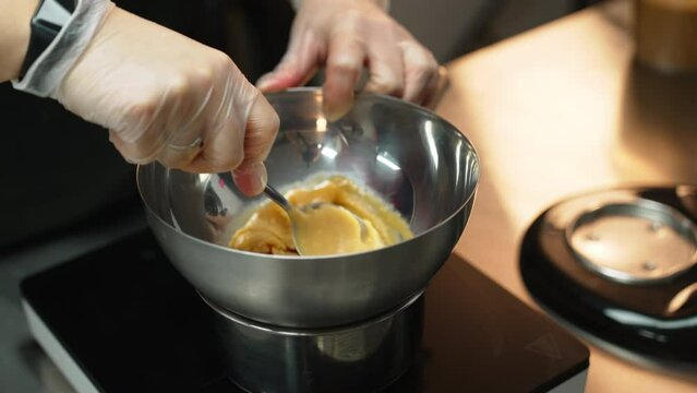 Baker wearing transparent gloves is mixing cream in metal bowl with tablespoon. Blurred background. High quality 4k footage