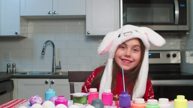 Cheerful little girl with easter bunny ears dances cheerfully in the kitchen and prepares for easter. Portrait of a cute cheerful girl joyfully dances and moves her Easter bunny ears kitchen table.