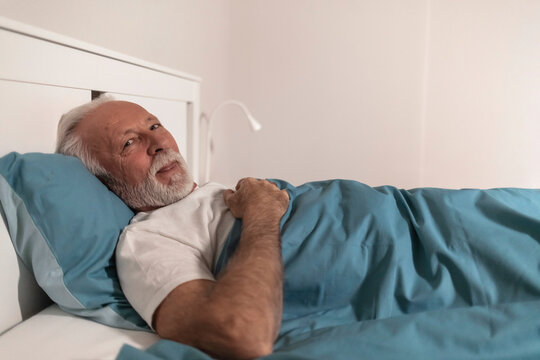 Senior Man In Bed In The Morning Smiling To The Camera. Closeup Front View Of Mid 60's Man Relaxing In Bed, Sincerely Smiling And Looking At Camera.