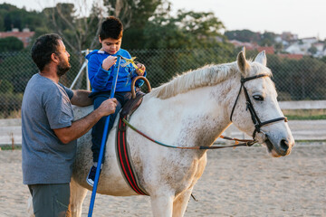 Child with disability doing exercises with the physiotherapist sitting on the horse outdoors.