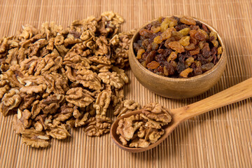 peeled walnuts are scattered on a wooden mat and in a wooden spoon and raisins in a wooden bowl nearby