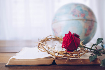 Close up of the red rose with the crown of thorns on an old open bible on wooden table with blurred world globe and white curtain background , Easter, love of God concept with copy space