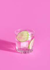 Minimal hot pink tropical fruit cocktail drink concept with ice and green lime in a crystal glass on girly pink background. Trendy sunlight summer party aesthetic.