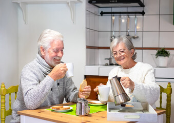 Smiling senior couple having breakfast together at the table at home. Elderly wife holding a coffee-maker, old people enjoying retirement