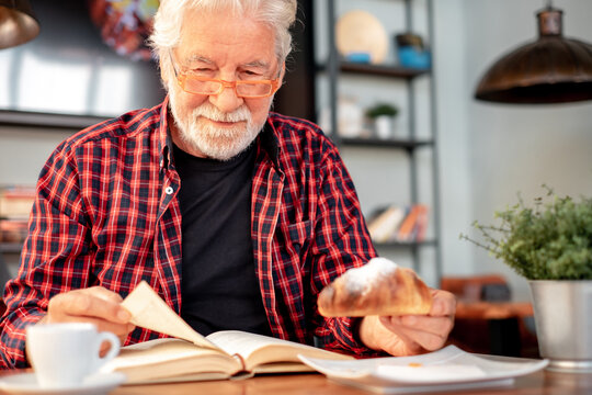 Smiling senior bearded man sitting at cafe table reading a book while enjoying breakfast with espresso coffee cup and croissant. Elderly caucasian male in eyeglasses and checkered shirt - Powered by Adobe