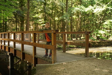A cute guy of 9-10-11 years old leans on the handrail of a wooden bridge in the forest and looks into the camera. Children's independence, children's loneliness