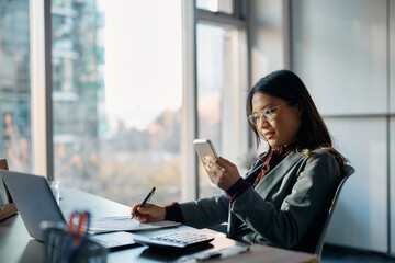 Smiling Asian businesswoman text messaging on cell phone while working in office.