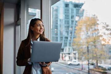 Happy businesswoman using laptop while looking through window in office.