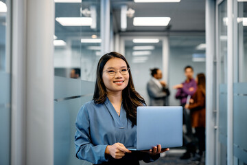 Happy Asian female entrepreneur with laptop in hallway looking at camera.
