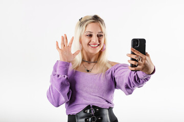 Young blonde girl smiling happy isolated over white background making a video call with smartphone and waving palm hand. Talks remotely with boyfriend or family. Laughs. Doing selfie.