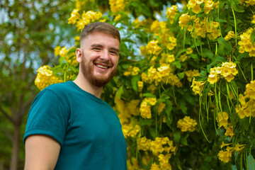 Naklejka na ściany i meble handsome young man smiling against the background of yellow blooming flowers in spring, summer time, healthy from allergies concept spring, blooming flowers, allergy, health