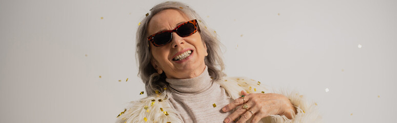 smiling senior woman in white faux fur jacket and trendy sunglasses near falling confetti on grey background, banner