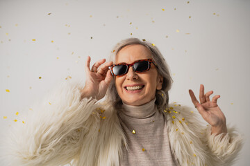 cheerful senior woman in white faux fur jacket adjusting trendy sunglasses near falling confetti on...