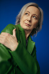 low angle view of thoughtful grey haired woman in green leather jacket looking away isolated on blue