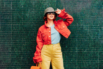 Hipster young woman in bright clothes, sun glasses, backpack bag and bucket hat posing on the green...