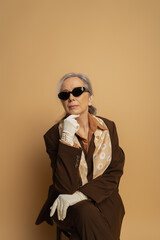 pensive senior woman in brown formal wear and sunglasses sitting on chair on beige